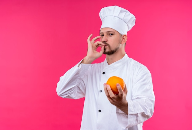 Satisfied professional male chef cook in white uniform and cook hat holding fresh orange fruit showing sign for delicious standing over pink background