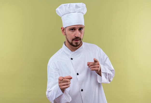 Satisfied professional male chef cook in white uniform and cook hat pointing with fingers to camera looking confident standing over green background