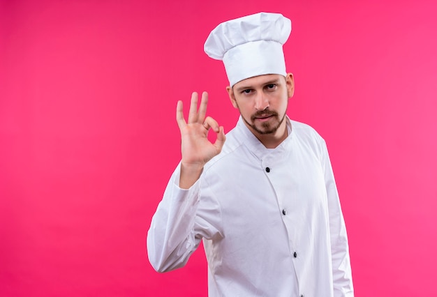 Satisfied professional male chef cook in white uniform and cook hat showing ok gesture looking confident standing over pink background