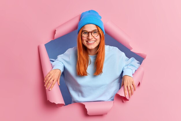 Satisfied redhead European woman smiles pleasantly has white teeth and freckled skin wears blue hat spectacles and sweatshirt stands in ripped hole of pink paper