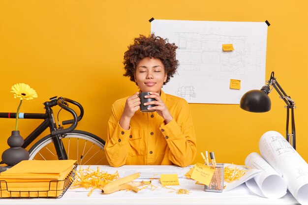 Satisfied relaxed woman architect office worker has coffee break closes eyes from pleasure as smells aromantic beverage draws draft at desktop creates sketches 