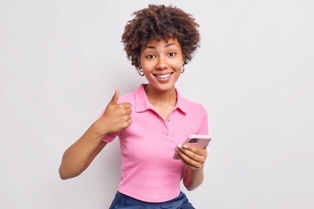 Satisfied woman holds mobile phone makes like gesture keeps thumb up says yes and recommends new application dressed in casual clothes isolated over white wall