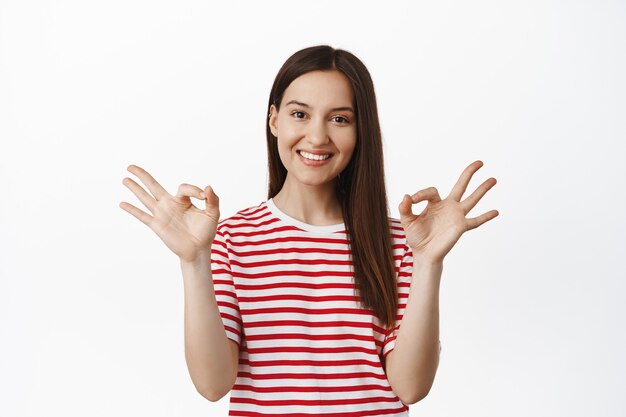 Satisfied young woman shows okay zero, OK sign and nod in approval, praise good choice, compliment you, agree and like something good, white wall.