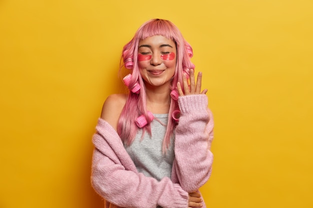 Satisified smiling woman with pink hair, applies rollers and beauty pads, dressed in rosy sweater, enjoys spare time for spending on herself