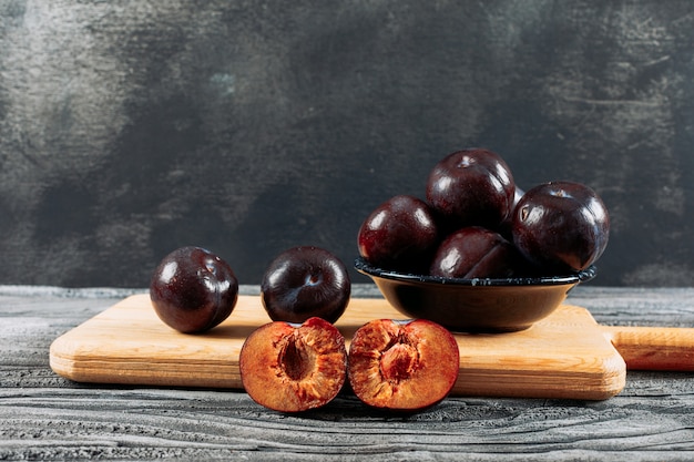 Savory plums in a dish and wooden board on a white wood and dark grey background. side view.