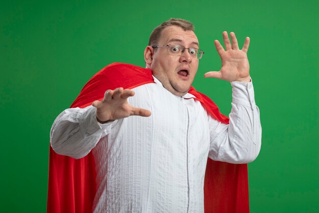 Scared adult slavic superhero man in red cape wearing glasses keeping hands in air looking at camera isolated on green background