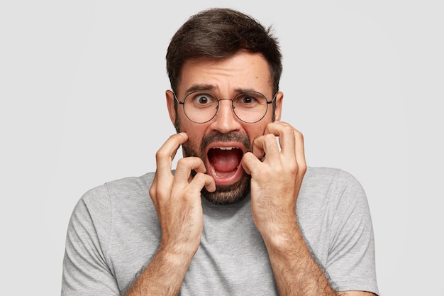 Free photo scared annoyed male opens mouth and frowns face, keeps hands on cheeks, looks desperately, being in panic, wears casual grey clothes, stands alone against white wall. negative feelings