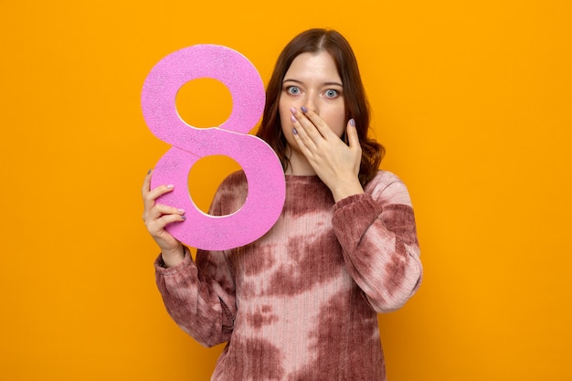 Scared covered mouth with hand beautiful young girl on happy women's day holding number eight 