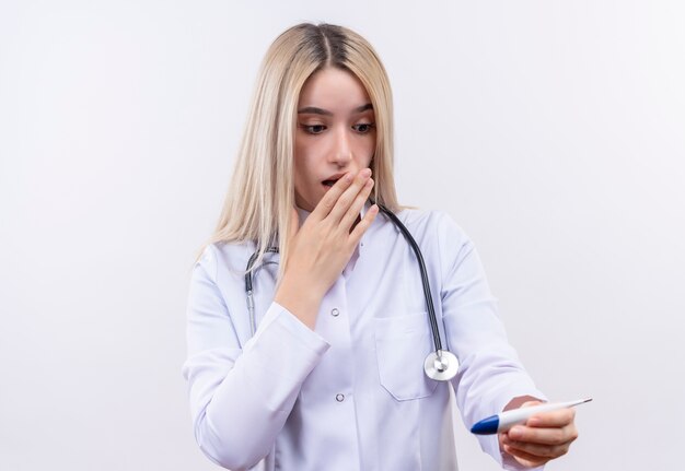 Scared doctor young blonde girl wearing stethoscope and medical gown looking at thermometer on hand covered mouth on isolated white background