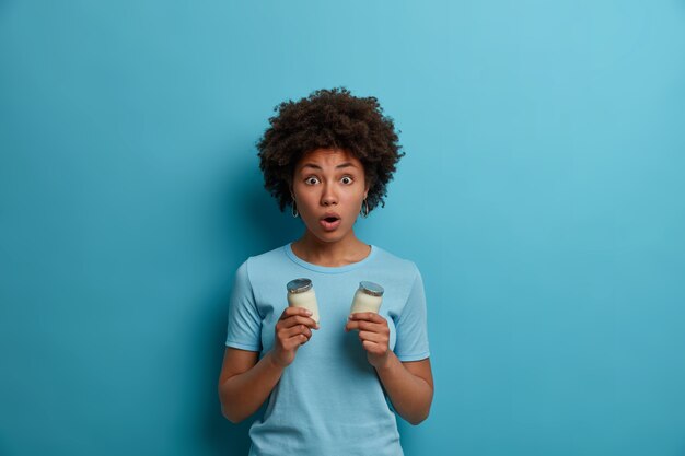 Scared emotional dark skinned woman gasps from shock, keeps jaw dropped, holds two glasses of yoghurt has healthy nutrition keeps to healthy diet wears casual blue t shirt. People, emotions, lifestyle