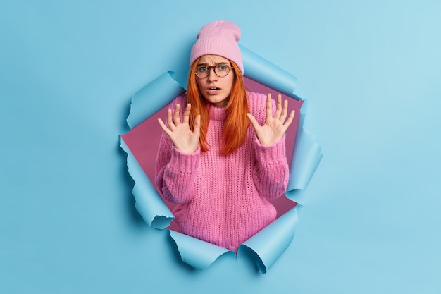 Free photo scared fearful woman raises hands in defensive gesture sees something horrible wears pink hat and sweater breaks through paper expresses negative emotions.