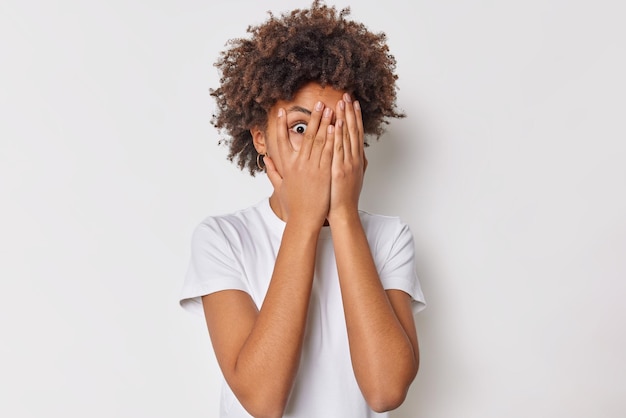 Scared frightened woman peeks through fingers covers face with hands feels danger panicking and trembles from fear dressed in casual t shirt isolated over white background. Human reaction concept