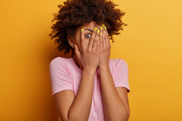 Scared frightened young woman peeks through fingers, hides face, stares with widely opened eyes at something spooky, wears casual t shirt, stands alarmed against yellow wall, being troubled