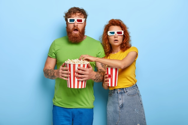 Free photo scared ginger couple watch scarying movie, gaze with stupefaction, eat popcorn, wear stereo glasses