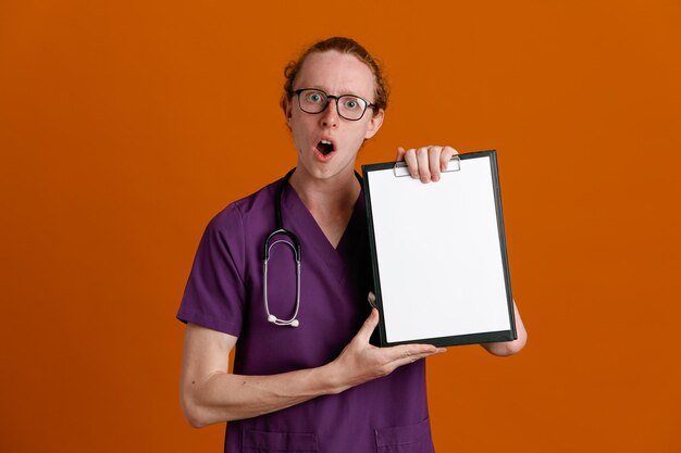 Scared holding clipboard young male doctor wearing uniform with stethoscope isolated on orange background