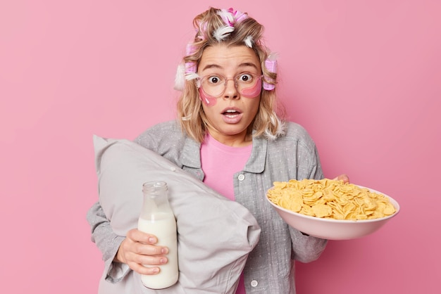 Free Photo scared impressed woman with hair rollers and feathers dressed in slumber suit holds pillow bowl of cornflakes and bottle of milk applies hydrogel patches under eyes isolated on pink background