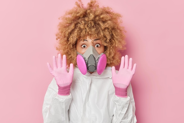 Free photo scared impressed young woman keeps palms raised towards camera tries to protect herself from danger dressed in protective white chemical suit gloves and gas mask isolated over pink background