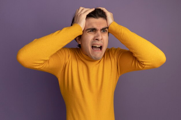 Scared looking at side young handsome guy wearing yellow turtleneck sweater grabbed head isolated on purple wall