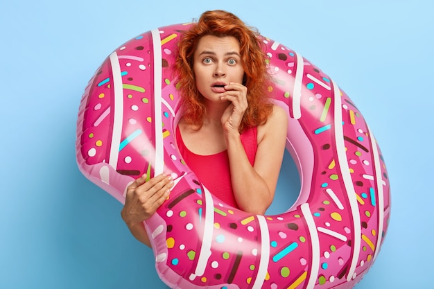 Scared lovely female model stares at camera, holds inflated swim ring, dressed in red bathing suit, shocked with spoiled summer vacation