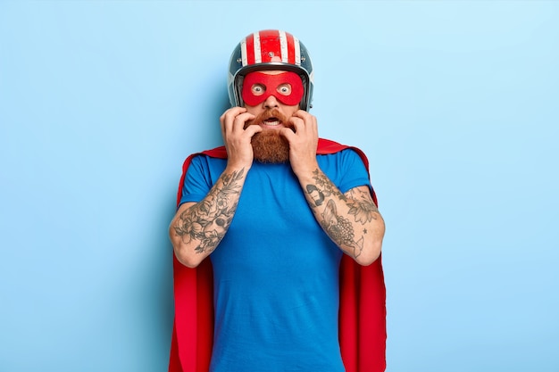 Free Photo scared nervous man looks with fear, wears helmet, red mask and cloak, prepares for flight