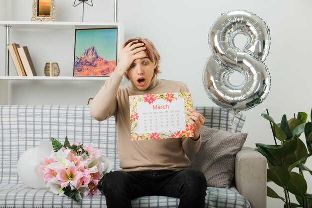 Scared putting hand on forehead handsome guy on happy women day holding calendar sitting on sofa in living room