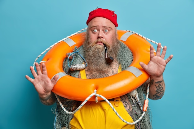 Free Photo scared seaman raises palm, stares bugged eyes, afraids of smimming, poses with orange inflated lifebuoy, fishing net, being seasick