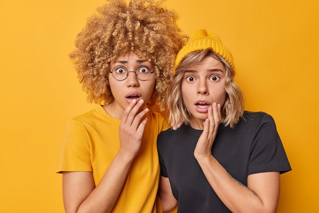 Scared shocked women stare with anxiety notice something awful and scarying dressed in casual t shirts stand closely to each other isolated over vivid yellow background People reactions concept