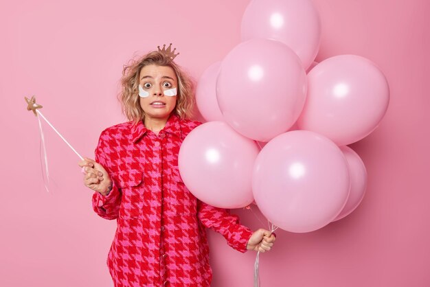 Scared terrified woman holds magic wand and bunch of inflated balloons cannot believe her eyes feels fearful applies beauty patches under eyes stands against pink background. Birthday party.