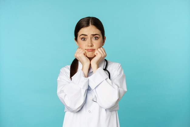 Scared and worried woman doctor in white coat, looking anxious and insecure, shaking from fear, standing over torquoise background.