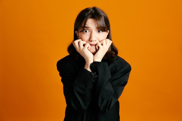 Scared young beautiful female wearing black jacket isolated on orange background