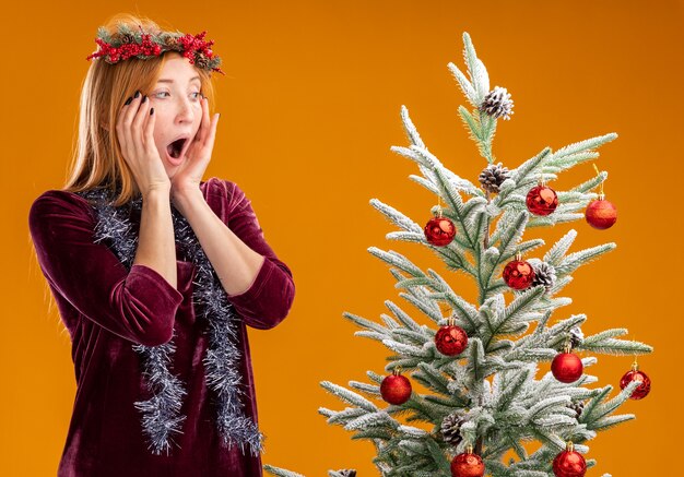 scared young beautiful girl standing nearby christmas tree wearing red dress and wreath with garland on neck looking at christmas tree putting hands on cheeks isolated on orange wall