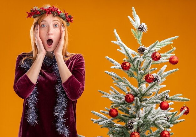 Scared young beautiful girl standing nearby christmas tree wearing red dress and wreath with garland on neck putting hands on cheeks isolated on orange background