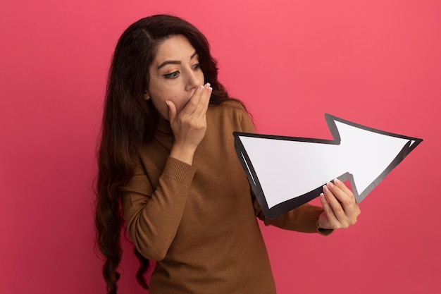 Free Photo scared young beautiful girl wearing turtleneck sweater holding and looking at direction mark isolated on pink wall