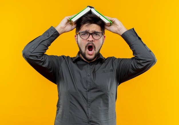 Scared young businessman wearing glasses covered head with book isolated on yellow