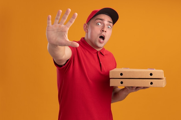 Scared young delivery man wearing uniform with cap holding pizza boxes showing stop gesture isolated on orange wall