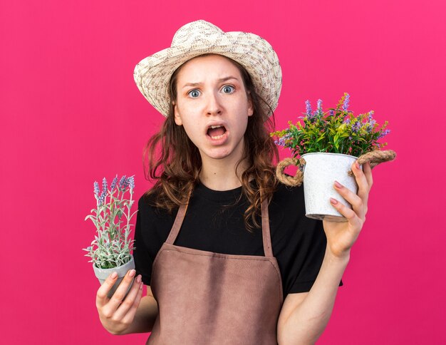 Scared young female gardener wearing gardening hat holding flowers in flowerpots 