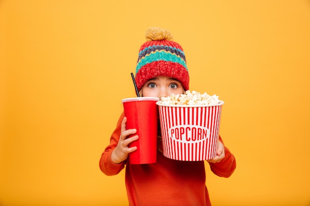 Free photo scared young girl in sweater and hat hiding behind the popcorn and plastic cup while looking at the camera over orange