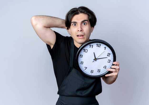 Scared young handsome male barber in uniform holding wall clock and putting hand on behind head isolated on white wall