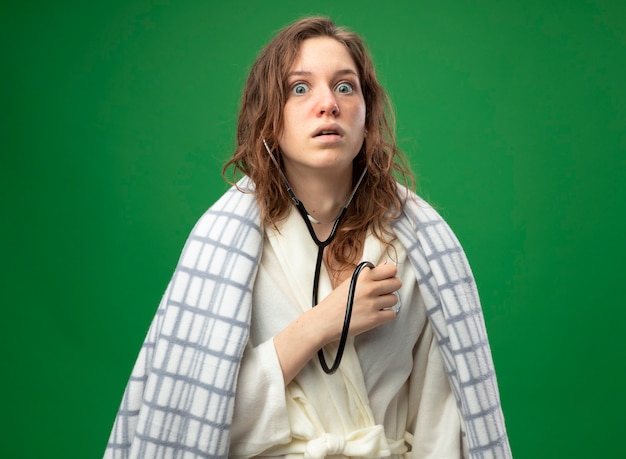 Free photo scared young ill girl wearing white robe wrapped in plaid listening to her own heartbeat with stethoscope isolated on green