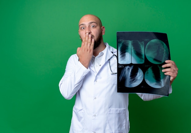 Free photo scared young male doctor wearing medical robe and stethoscope holding x-ray and covered mouth with hand isolated on green background