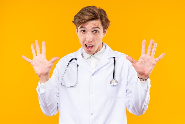 scared young male doctor wearing medical robe with stethoscope showing stop gesture isolated on orange wall