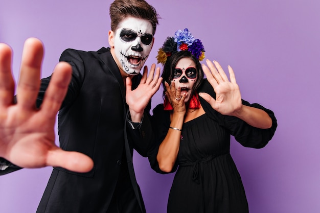 Free photo scared young people in halloween attires standing together on purple background. indoor photo of enthusiastic european couple fooling around in muertos costumes.