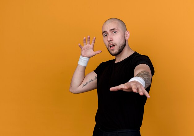 Scared young sporty man wearing wristband holding out hand isolated on orange wall with copy space