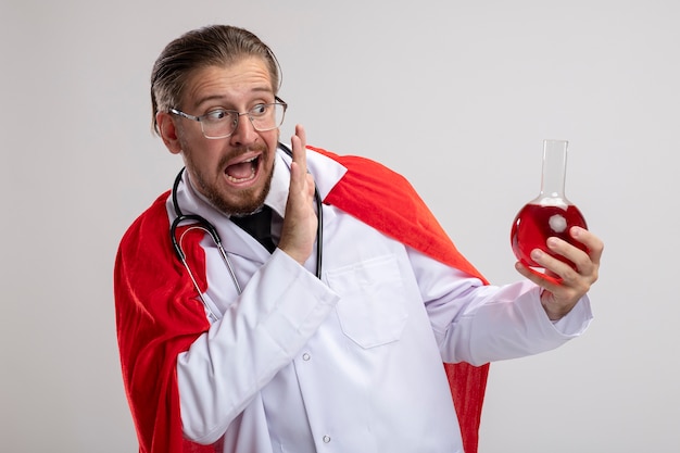 Scared young superhero guy wearing medical robe with stethoscope and glasses holding chemistry glass bottle filled with red liquid isolated on white background