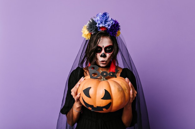 Free Photo scary dead bride holding pumpkin. european woman in black veil posing on purple wall in halloween.