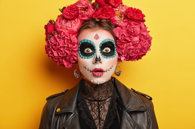 Scary woman wears horror Halloween makeup, has frightened expression, chas dark painted circles around eyes, wears big red flower wreath, isolated over yellow background.