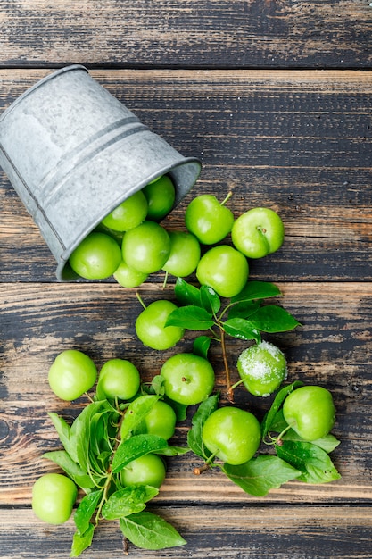 Scattered green plums with leaves, salt from a mini bucket on wooden wall, top view.