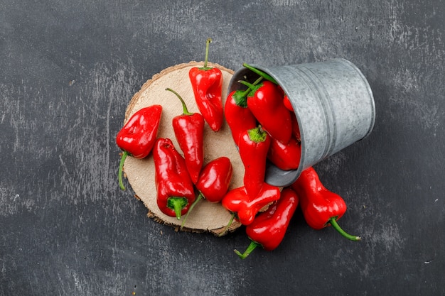 Scattered red peppers from a mini bucket with wooden piece top view on a grungy grey wall