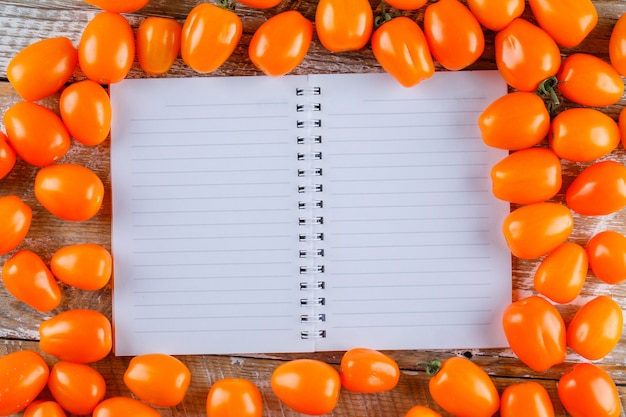 Scattered tomatoes with opened copybook on wooden table, flat lay.