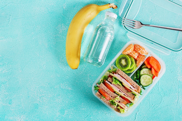 Free photo school lunch box with sandwich, vegetables, water, and fruits on table